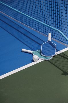 a tennis racket and ball laying on the ground in front of a blue net