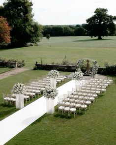 an outdoor ceremony set up with white chairs and floral arrangements on the aisle, in front of a large field