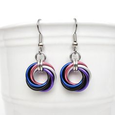 Chainmail love knot earrings are simple and understated earrings that are great for work or play. Lightweight anodized aluminum jump rings in the genderfluid pride flag colors of pink, white (represented here with matte silver), purple, black, and blue were handwoven together to create these earrings. The swirl of the knot is created when each new open jump ring is passed through all previous rings and then closed.  These earrings measure 1-1/2 inch including the hypoallergenic stainless steel ear wires. They are 5/8 inch wide. Dime shown for scale. The current style of ear wires are stainless steel French hooks, but can be switched to stainless lever backs if you prefer. Please select your choice from the drop down menu.  Aluminum is a lightweight metal that will not tarnish or rust, and Pride Earrings, Genderfluid Pride, Pride Flag Colors, Pride Jewellery, Chainmail Jewelry, Chain Maille Jewelry, White Jewelry Box, Chain Maille, Hard Metal