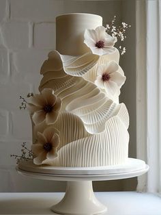 a three tiered cake with white icing and flowers on top, sitting in front of a window