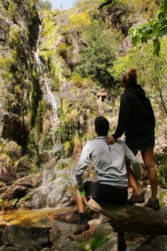 two people sitting on a log looking at a waterfall