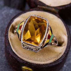 This fun ring features a checkerboard rectangular cut citrine flanked by yellow, black, green, and red enamel accented shoulders. The ring is crafted in 10k white gold and is currently a size 5.5. Due to the enamel details, resizing options are limited. Luxury Vintage Gold Enamel Ring, Art Deco Enamel Ring As A Gift, Art Deco Enamel Ring As Gift, Art Deco Enamel Ring Jewelry, Art Deco Enamel Jewelry Ring, Luxury Yellow Rings For Collectors, Collectible Hallmarked Enamel Ring In Fine Jewelry Style, Vintage Yellow Enamel Jewelry, Enamel Multi-stone Rings As A Gift