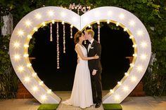 a bride and groom kissing in front of a heart shaped sign with lights on it