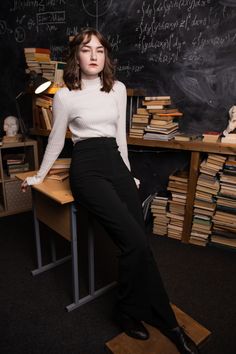 a woman sitting on top of a wooden desk in front of a blackboard filled with books