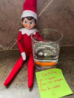 an elf sitting next to a glass filled with liquid and a note on the floor