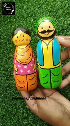 two small wooden dolls sitting on top of green grass in front of a person's hand