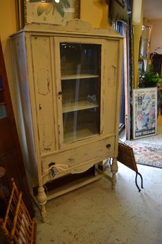 an old white china cabinet with glass doors in a living room next to a chair