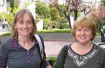 two women standing next to each other in front of trees and bushes on the street