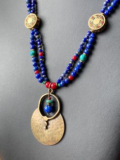 Featuring stones that are associated with wisdom, insight, spirituality, and communication, this is made with gorgeous deep royal blue to navy blue grade A Lapis in various shapes and sizes, which contrast strikingly with green semi precious turquoise, rich red Czech glass and white-heart beads, and brass spacers. The two strands gather at Tibetan brass 15mm coin beads inlaid with red coral and turquoise howlite as well as other brass beads. A 53x37mm plated antique gold large tribal style medal Bohemian Blue Lapis Lazuli Beaded Necklace, Blue Lapis Lazuli Jewelry For Festivals, Handmade Bohemian Sapphire Necklace, Bohemian Lapis Lazuli Beaded Necklaces As Gift, Bohemian Lapis Lazuli Beaded Necklaces For Gift, Blue Amulet Necklaces With Natural Stones, Blue Amulet Style Necklace With Natural Stones, Bohemian Lapis Lazuli Beaded Necklace For Gift, Blue Amulet Necklace With Natural Stones