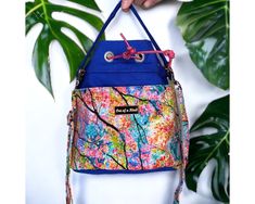 a hand holding a multicolored purse with flowers on the front and side, hanging from a plant