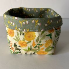 a yellow flowered fabric basket on a white surface with green trimming and flowers