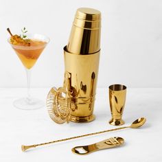 a gold cocktail shaker next to two glasses and spoons on a white surface