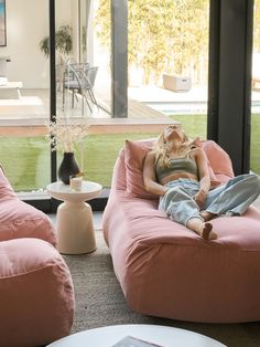 a woman sitting on a pink bean bag chair in front of a large window with grass outside