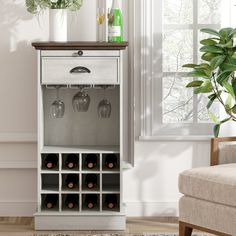 a white cabinet with wine glasses and bottles in it next to a chair near a window