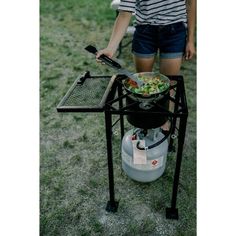 a woman standing next to a grill on top of a grass covered field holding a frying pan