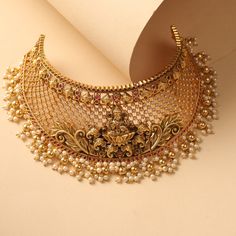 Description Inspired by the classic temple jewelry from the south, this antique choker necklace is crafted with the Goddess Laxmi and Ganesh motifs with the ivy design along with semi-precious CZ stones. With high quality craftsmanship and the fine finish, this piece is perfect to adorn in wedding and festivals. The guttapusalu drop pearls add the extra charm to this beautiful jewelry design. Details & Specifications: Materials used : Brass Alloy with Antique Gold Plating Weight – Choker 63.51 g South Temple Jewellery, South Indian Motifs, South Indian Gold Jewellery, Antique Gold Choker Set, Gold Choker Necklace Set, Antique Choker, Goddess Laxmi, Choker Necklace Designs, Perhiasan India