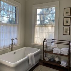 a white bath tub sitting under two windows next to a shelf with towels on it