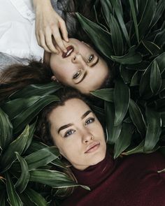 two women laying on the ground surrounded by green leaves and plants, one with her eyes closed