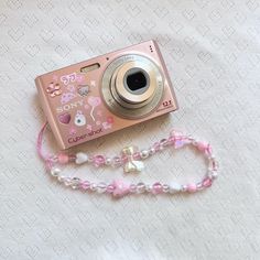 a pink camera sitting on top of a white bed next to a beaded necklace