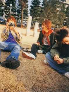 three people sitting on the ground in front of some trees and grass with one person taking a photo