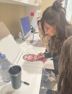 two women are working on drawings at a desk with water bottles and pens in front of them