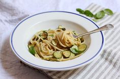 a white bowl filled with pasta and vegetables on top of a striped cloth next to a spoon