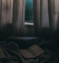 an open book sitting on top of a bed in front of a window with curtains