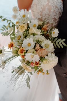 a bride and groom holding a bouquet of white and orange flowers in front of the ocean