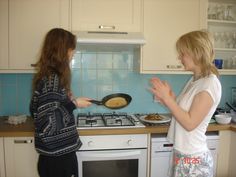 two women standing in a kitchen cooking food on top of a stove and one is holding her hands out