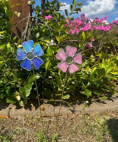 two stained glass flowers sitting in the middle of a flower bed with pink and blue flowers