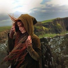 a woman with long hair standing in front of a stone wall and holding her hands to her face