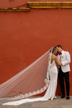 a bride and groom standing next to each other in front of a red wall with a white veil