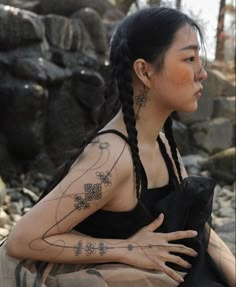 a woman with tattoos on her arms and chest sitting in front of rocks, looking off into the distance