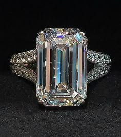 an emerald cut diamond ring with diamonds around it's shoulders and sides, on a black surface