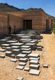 two people standing in front of a building made out of concrete blocks with mountains in the background