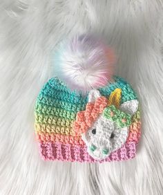 a crocheted hat with a rainbow unicorn on the front and a white pom - pom in the back