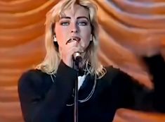 blond haired woman singing into a microphone in front of a stage curtain with curtains behind her