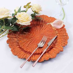 an orange plate with flowers and silverware on it next to a glass vase filled with white roses