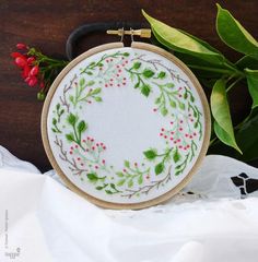 a cross stitch wreath with red berries and green leaves is on a white cloth next to a plant