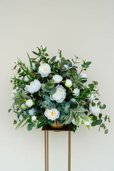 white flowers and greenery in a gold vase on a stand against a beige wall