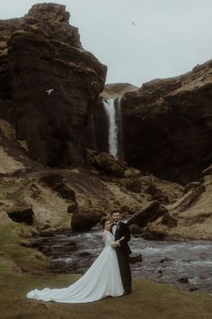 a bride and groom standing in front of a waterfall