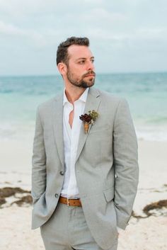 a man in a gray suit and white shirt standing on the beach with his hands in his pockets