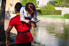 two people dressed in red and black are looking at a book while standing next to a lake