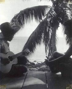 an old photo of a man playing the guitar under a palm tree by the ocean