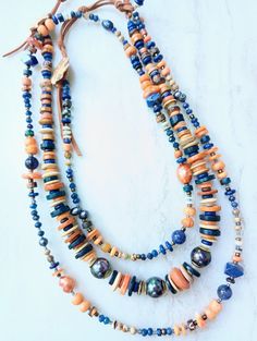 Two bright, bold necklaces. An array of oranges, rusts, blues, beige, white, and navy. Many different shapes and finishes. Both necklaces are full of Lapis, Orange Howlite, Jasper, Picture Jasper, dyed Turquoise in coral color, stunning coral beads, Mother of Pearl nuggets, colorful pearls, and more. Mix in bone beads, Java Glass in burnt orange and a mixed stripe, shell disks, mixed metals, dyed coconut wood disks, and more. Both creations are strung on Beadalon .018 7-strand Stainless Steel Be Orange Bohemian Necklace, Handmade Orange Multi-strand Necklace, Earthy Handmade Blue Necklace, Colorful Pearls, Coconut Wood, Chunky Statement Necklace, Wood Disc, Bold Necklace, Bone Beads