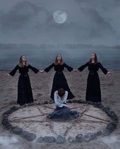 four women in long dresses are standing on the sand with their hands out to each other