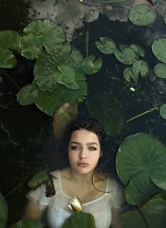 a woman is standing in the water surrounded by lily pads and green leaves, looking up at the camera