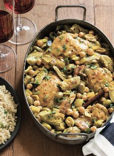 a pan filled with chicken, beans and rice next to two glasses of red wine