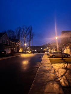 an empty street at night with cars parked on the side and houses in the background