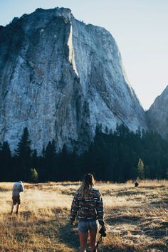 a woman walking through a field next to a large rock face in the background with trees and grass